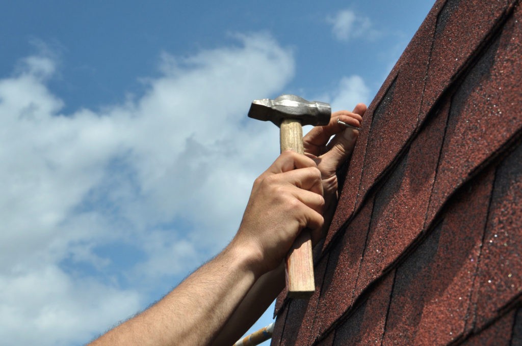 close up of man's hands hammering a nail to dark brown asphalt shingles and blue sky in the background