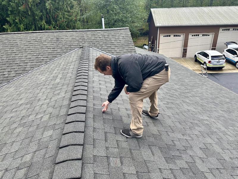 a man in a black jacket and beige trousers on a cobblestone 3-tab asphalt shingle roof, inspecting for granule loss.